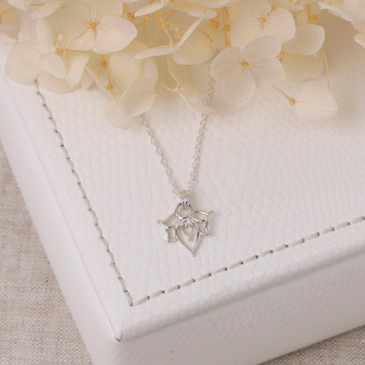Silver necklace with a lotus-shaped pendant on a white jewelry box with flowers in the background