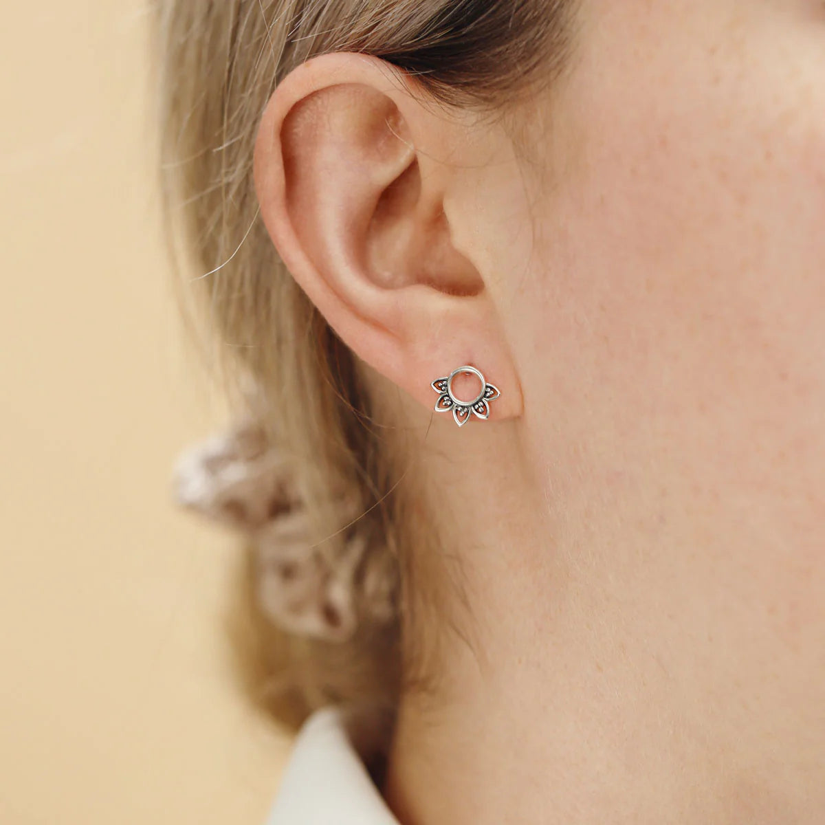 Close-up of an ear wearing a small silver earring with a circular rising sun design on a beige background.