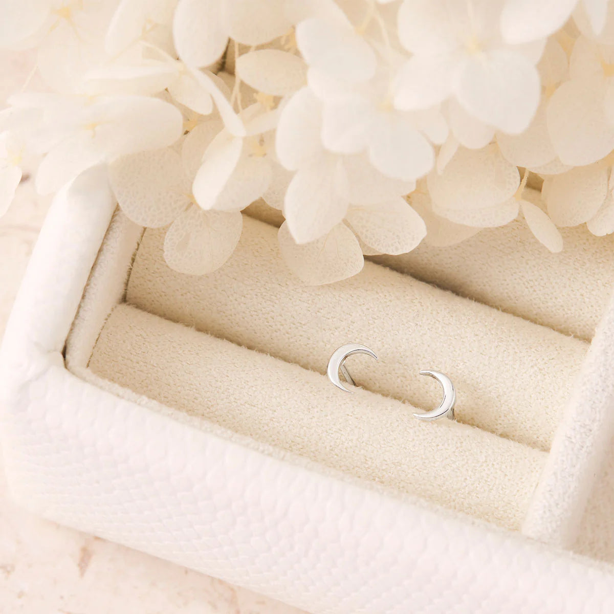 Silver cresent moon studs on a soft beige surface with light-colored flowers in the background