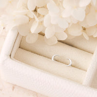 Silver cresent moon studs on a soft beige surface with light-colored flowers in the background