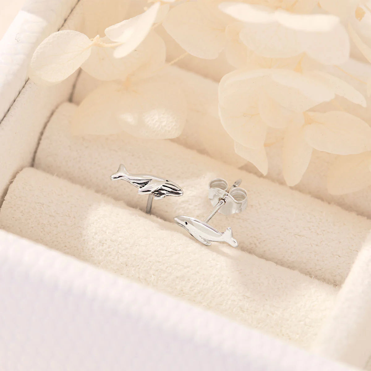 Silver whale-shaped earrings on a soft beige fabric background
