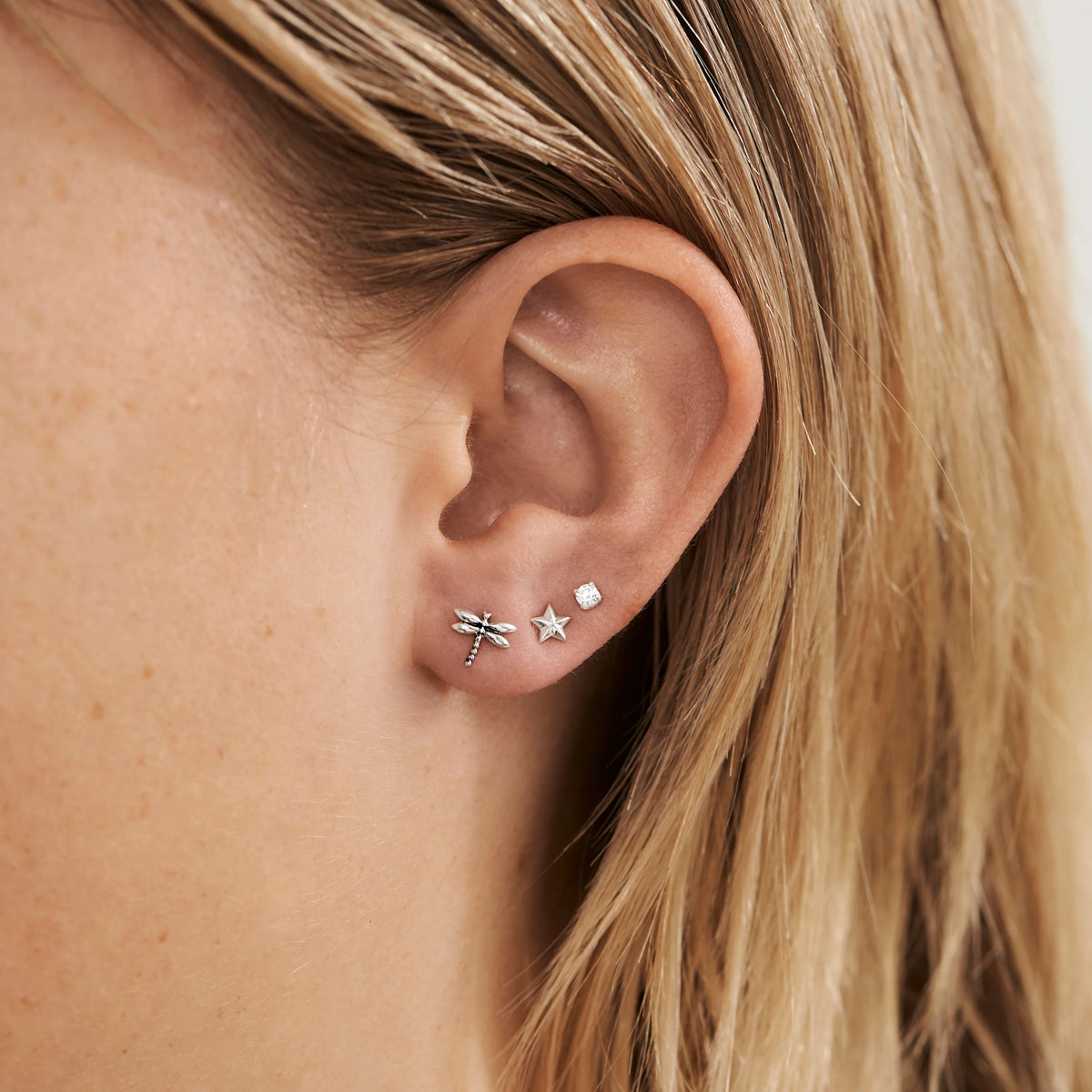 Close-up of an ear wearing three earrings: a star, a dragonfly, and a small round one.