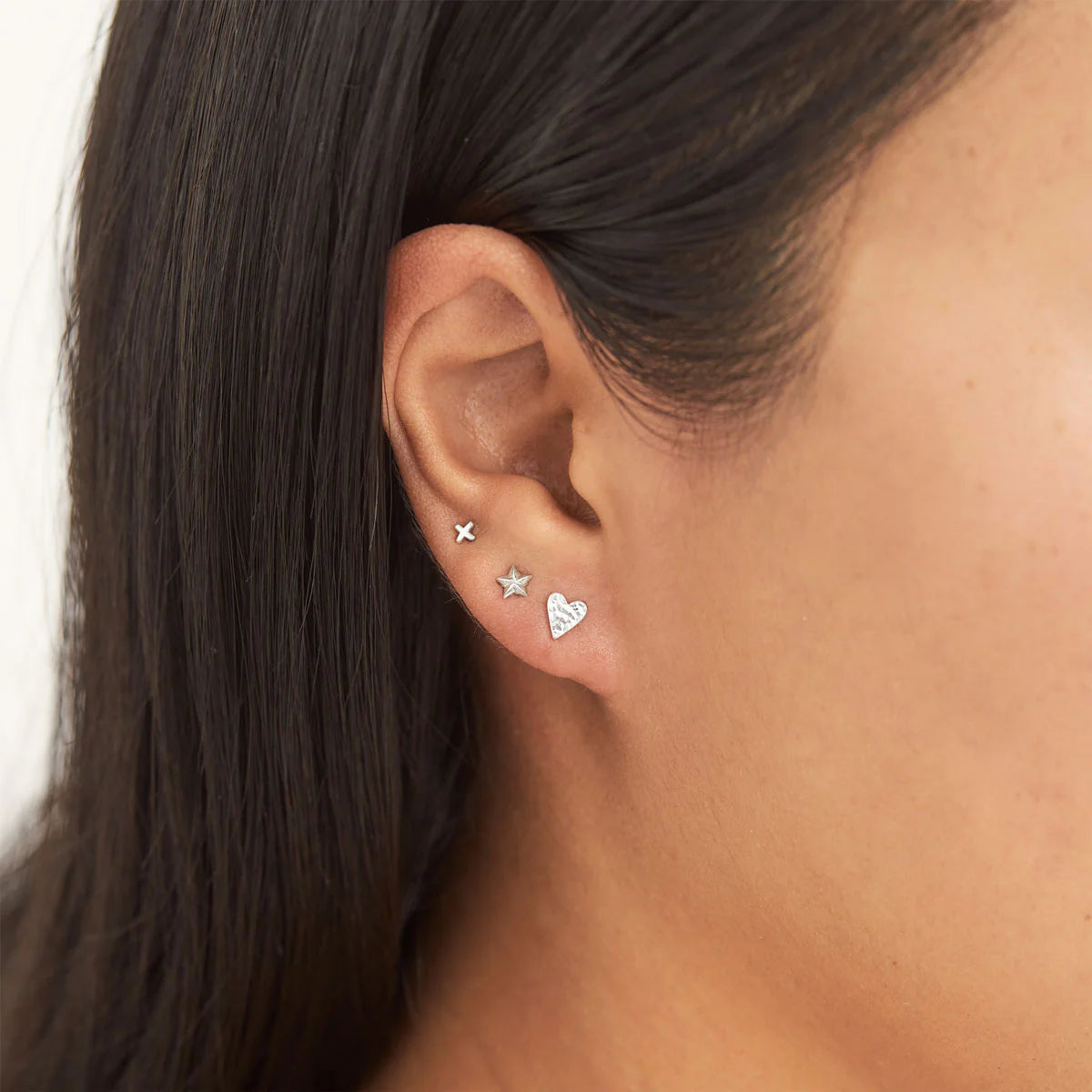 Close-up of an ear wearing three earrings: a star, a heart, and a cross.
